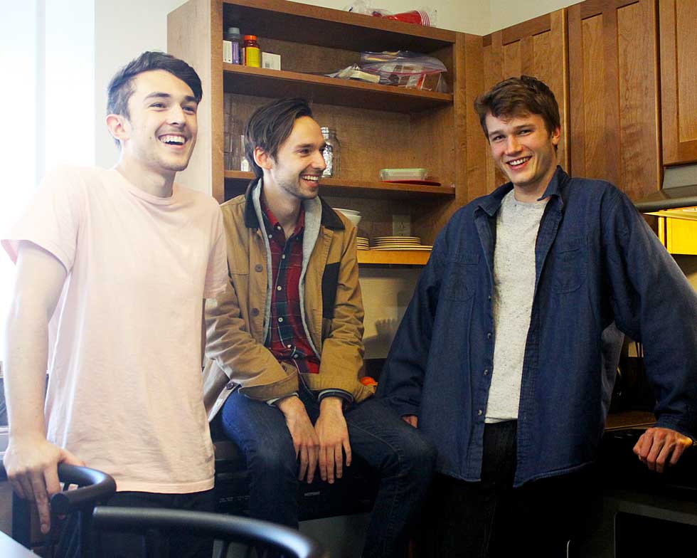 Three students smile and laugh together in a shared apartment kitchen with wooden cabinets and open shelving.