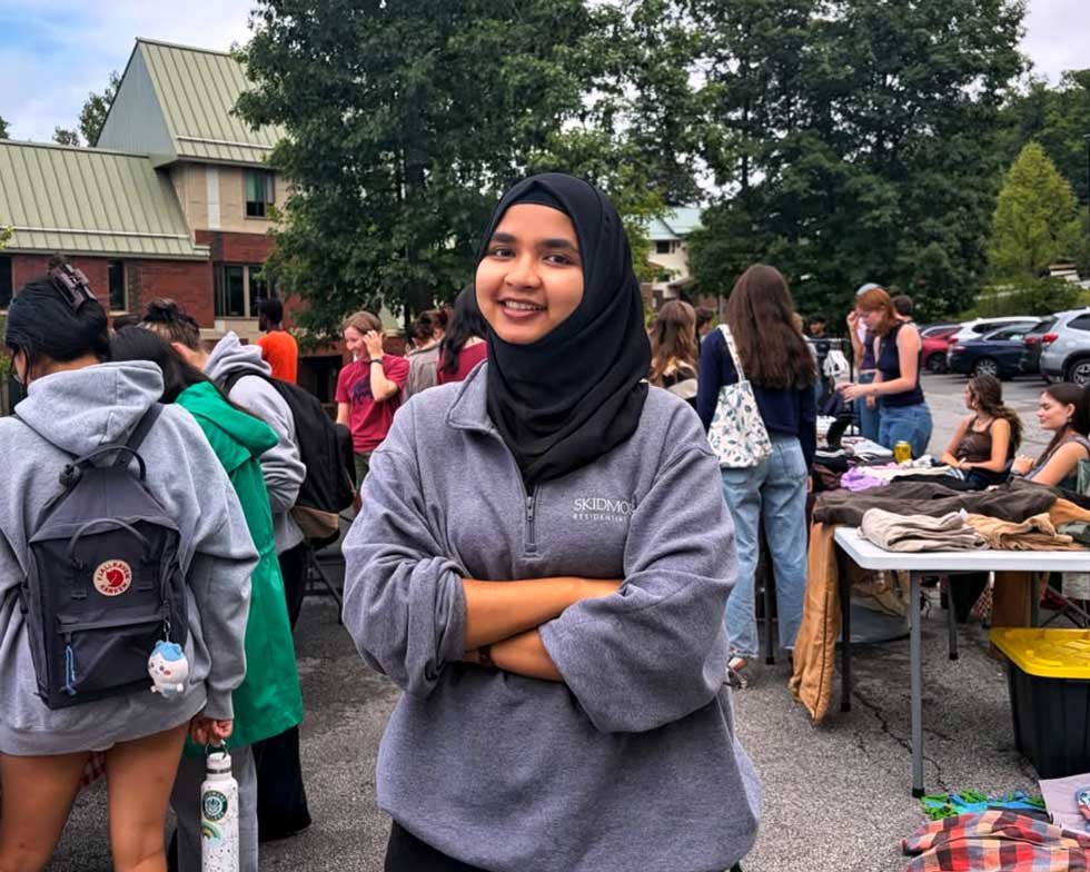 A smiling student in a Skidmore quarter-zip stands with arms crossed at an outdoor campus event with clothing tables and other students in the background.
