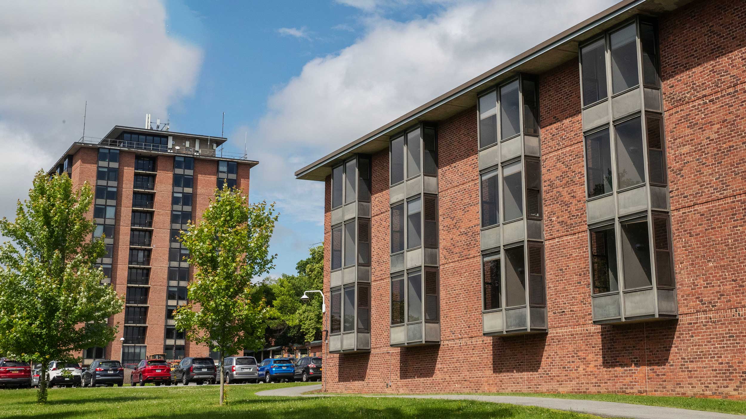 Brick Skidmore residence halls with tall windows, trees, parked cars, and a partly cloudy sky.