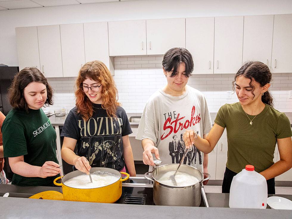 Four students cook together in a kitchen. Each are stirring from two different pots and there are white kitchen cabinets hanging behind them.