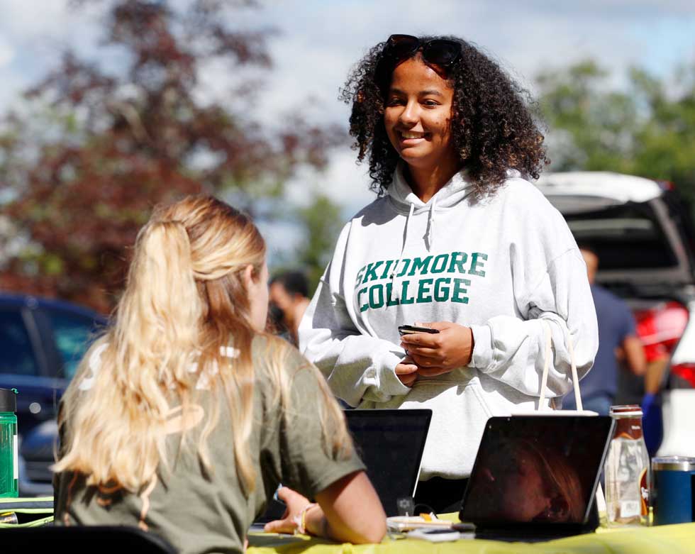 A student in a Skidmore College hoodie talks with another student at an outdoor residence life table.