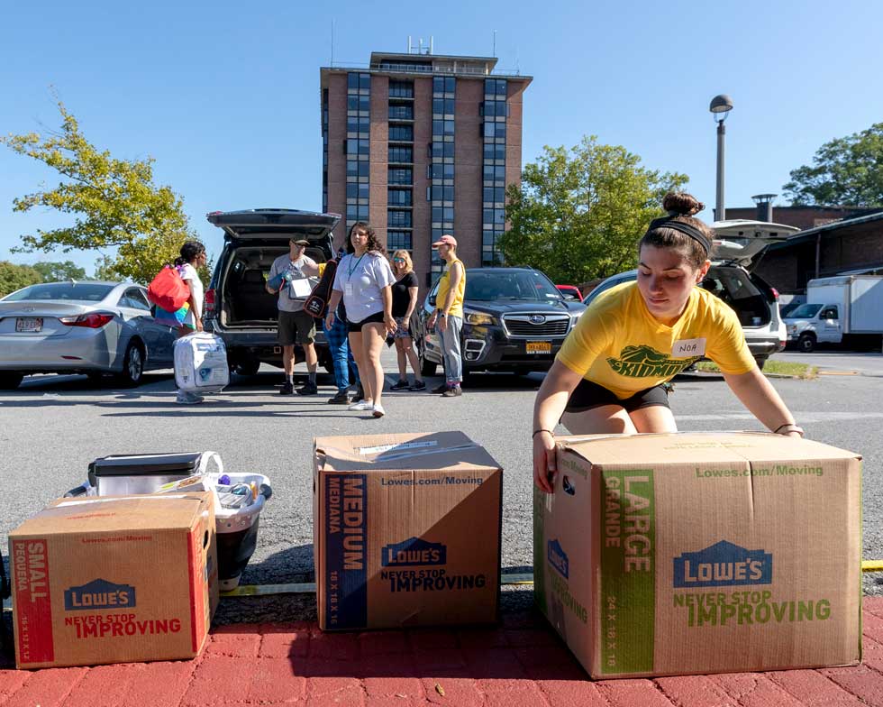 A student helps carry moving boxes during residence hall move-in, with families and cars in the background.