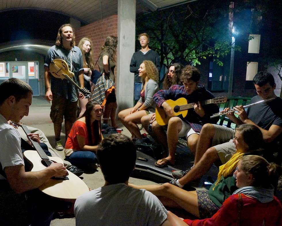 Students gather outdoors at night to sing and play guitars, mandolin, and flute near a residence hall.
