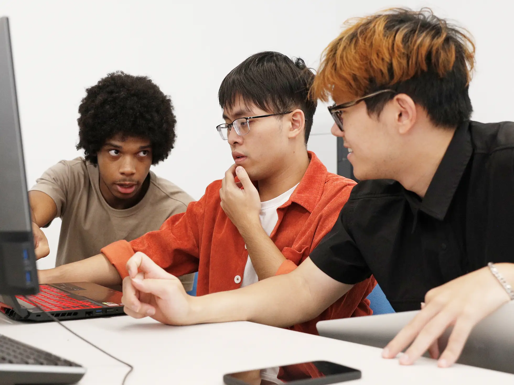 Three computer science college students work together intently at a desk with laptops, one pointing at a screen while the others focus on the task.