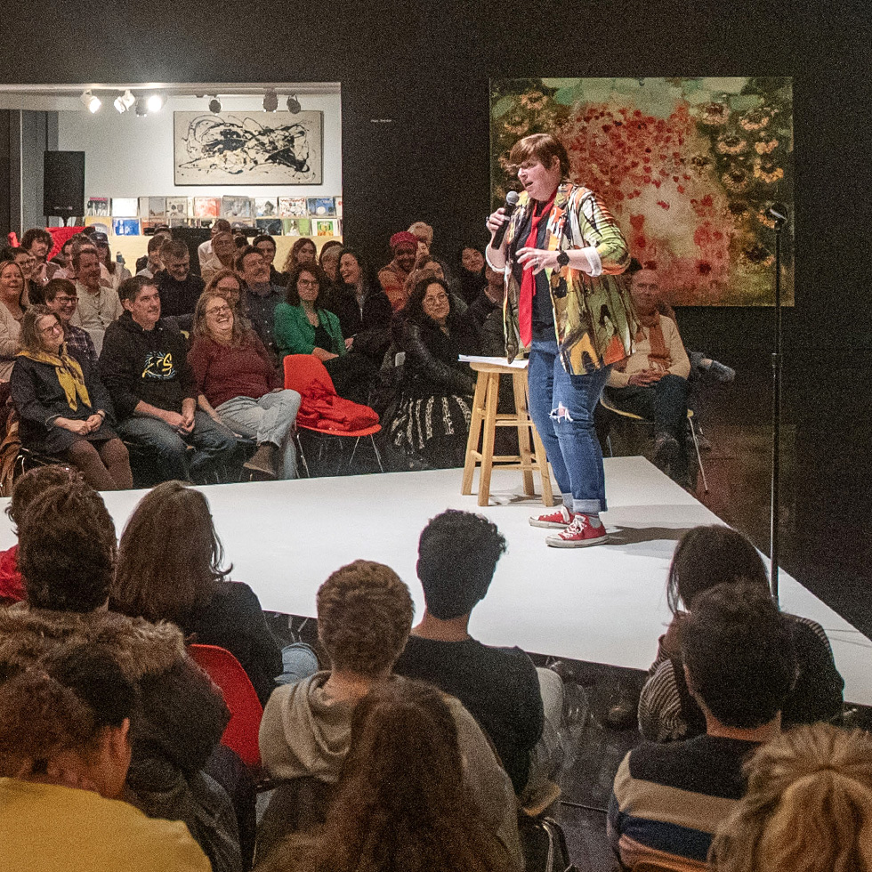 A professor stands on a small stage with a microphone, entertaining a packed audience in an art gallery with colorful artwork in the background.
