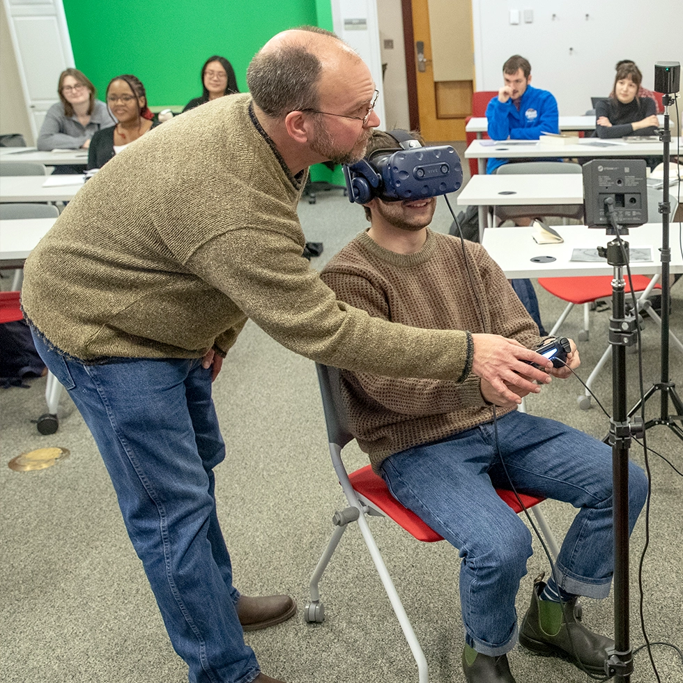 A professor assists a student wearing a VR headset and holding a controller, while other students watch from their desks in a technology-equipped classroom.