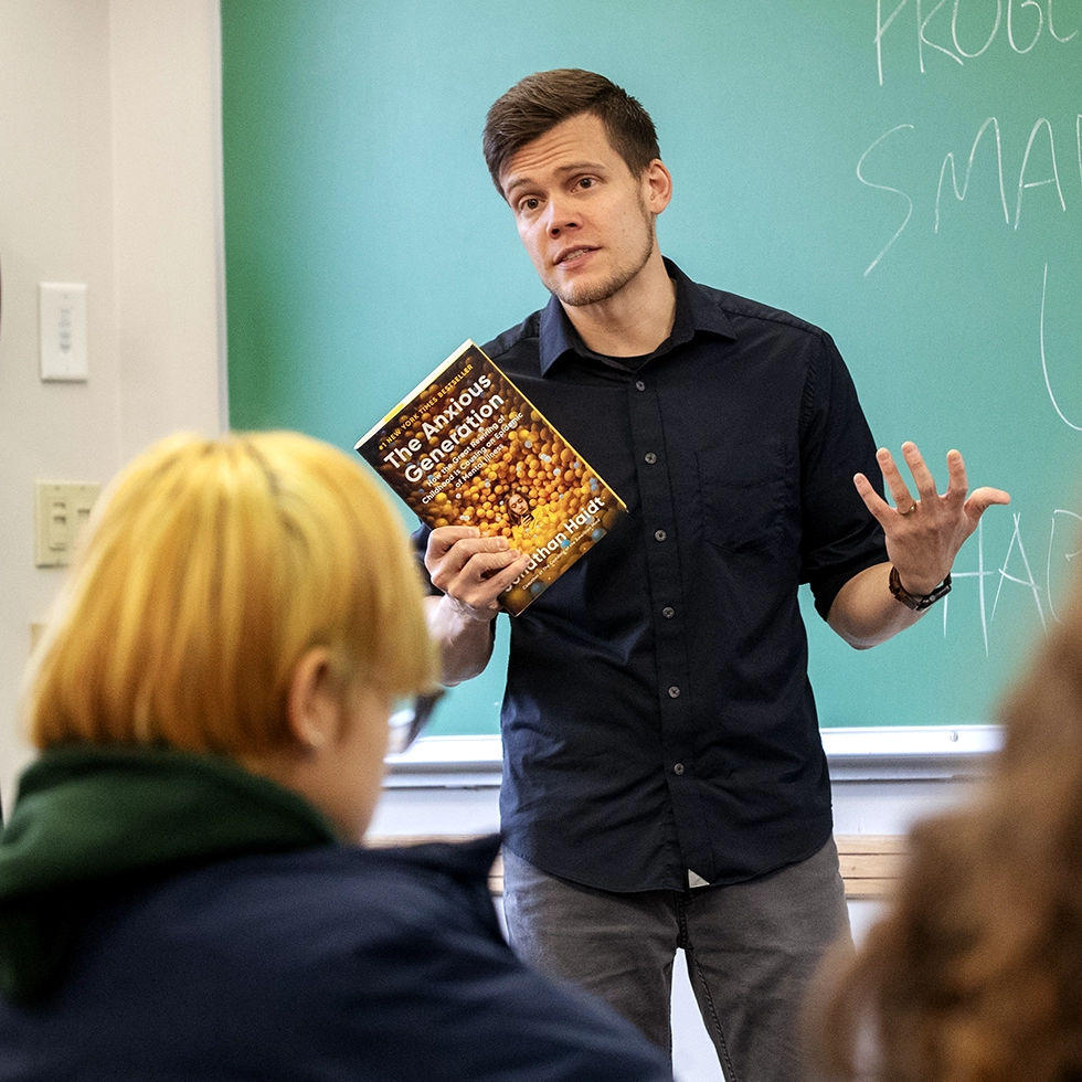 A professor holds up a copy of The Anxious Generation while addressing students in a classroom with a green chalkboard.