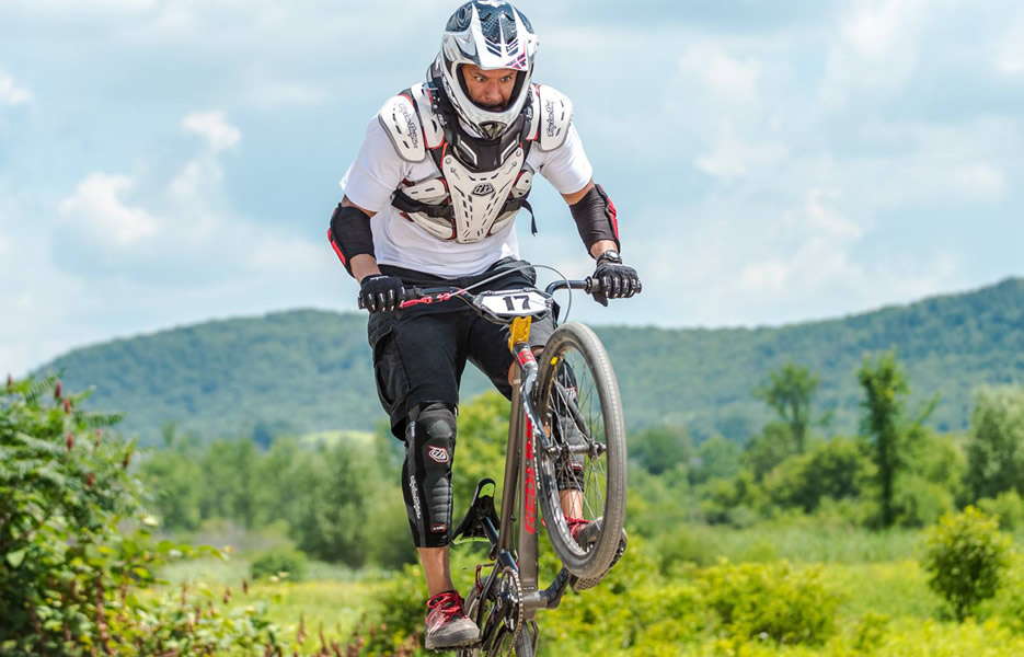 A mountain biker wearing a helmet and protective gear lifts off the ground on a dirt trail, with lush green hills and trees in the background.