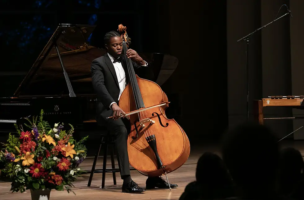 A Skidmore student in formal attire performs a solo on double bass in a recital hall, with a grand piano and floral arrangement nearby.