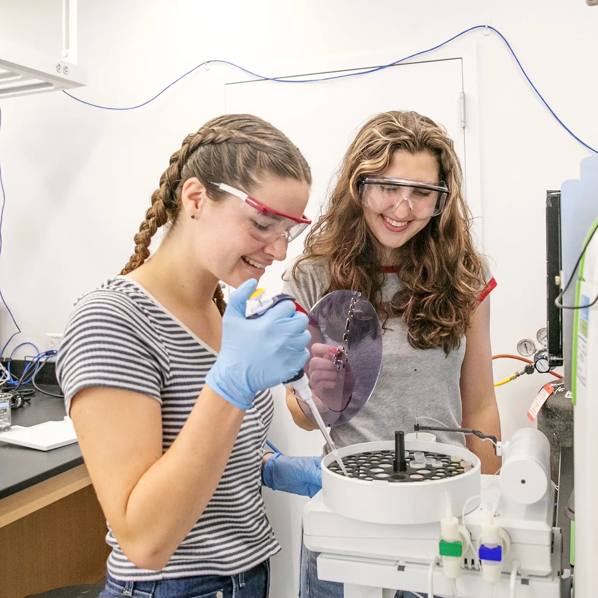 Two students in safety goggles and gloves conduct a chemistry experiment in a Skidmore science lab, smiling as they work together