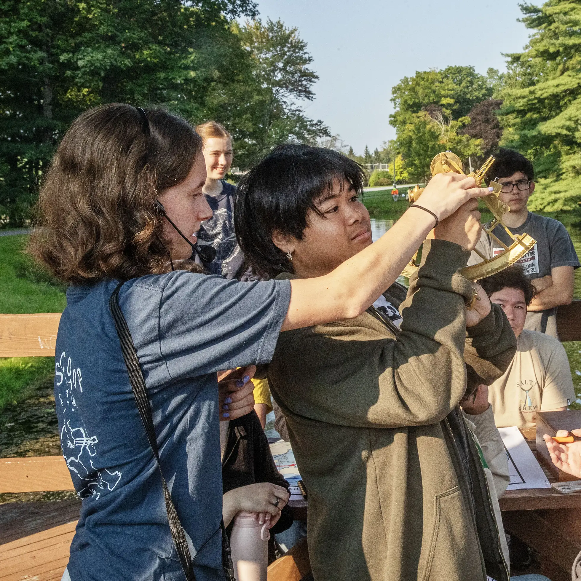 Students in an outdoor classroom at Skidmore use historical navigation tools, including a brass sextant, during a hands-on learning activity by the water.