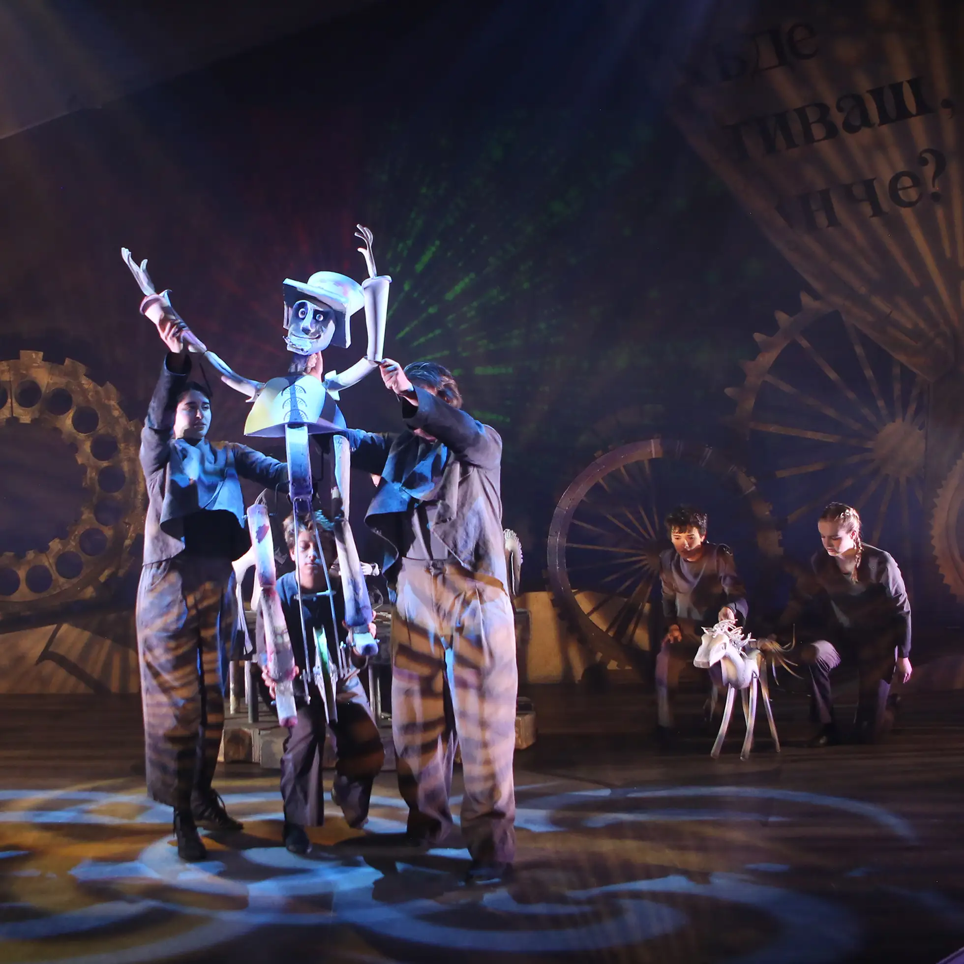 students on stage manipulate large-scale puppets during a theatrical performance, surrounded by dramatic lighting and oversized gears.