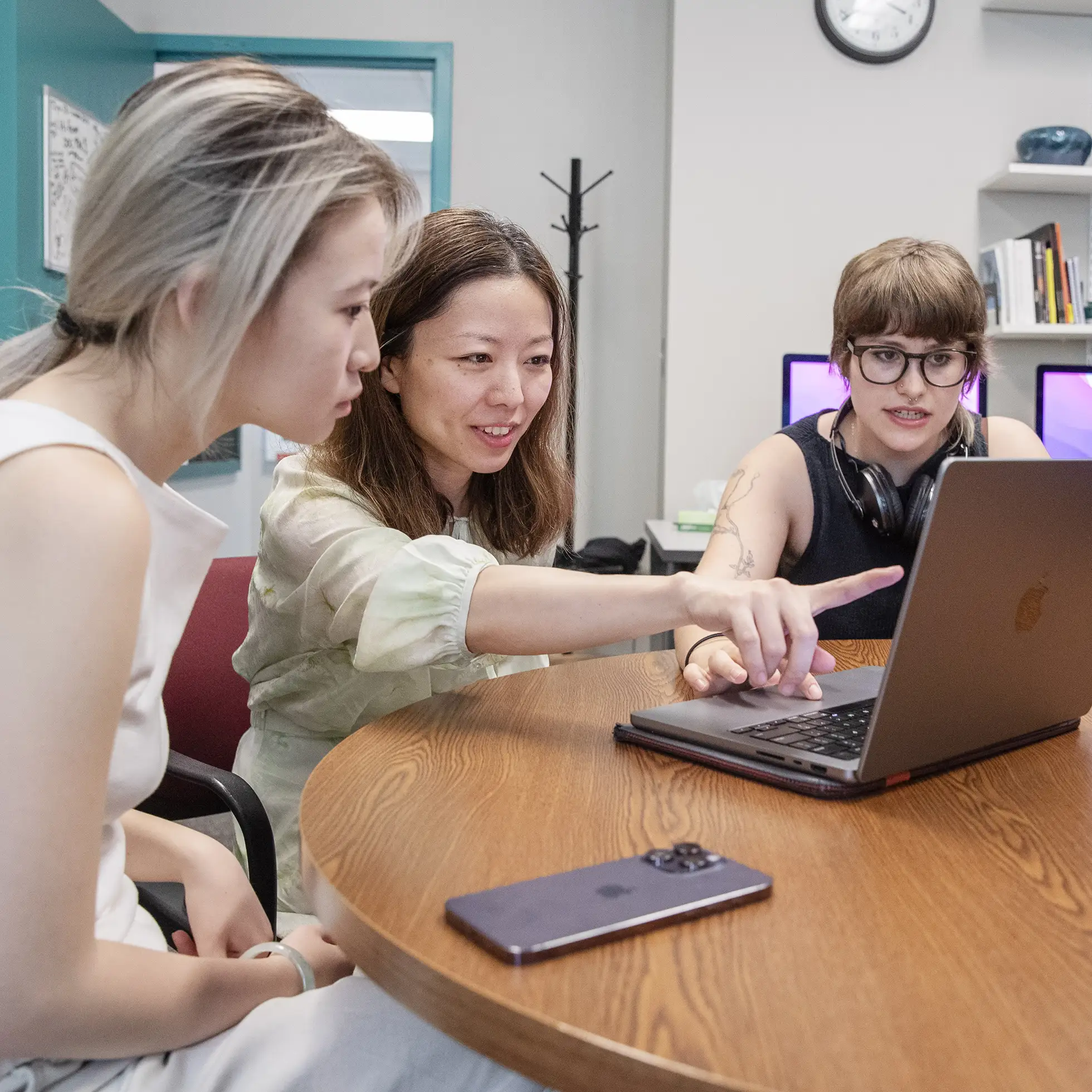 A professor and two students collaborate around a laptop in a classroom setting.