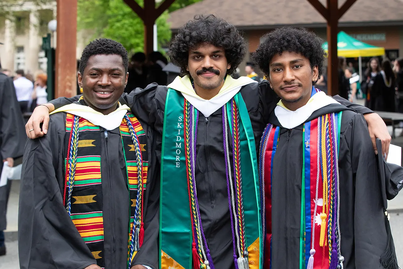 Three graduating students in caps and gowns stand arm-in-arm, smiling and wearing colorful stoles and cords that represent academic honors, cultural heritage, and campus involvement during Skidmore College’s Commencement ceremony.