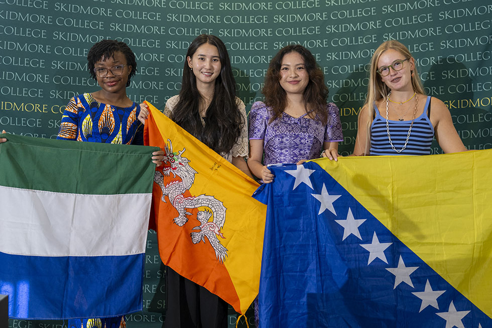 Four international students at Skidmore College stand in front of a green Skidmore College backdrop, each holding a national flag. From left to right, the flags are from Sierra Leone, Bhutan, Bosnia and Herzegovina.