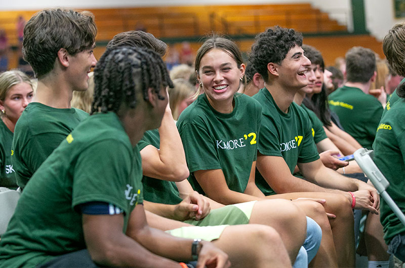 Students wearing matching green shirts that says "Skidmore" great each other while seated in a gymnasium