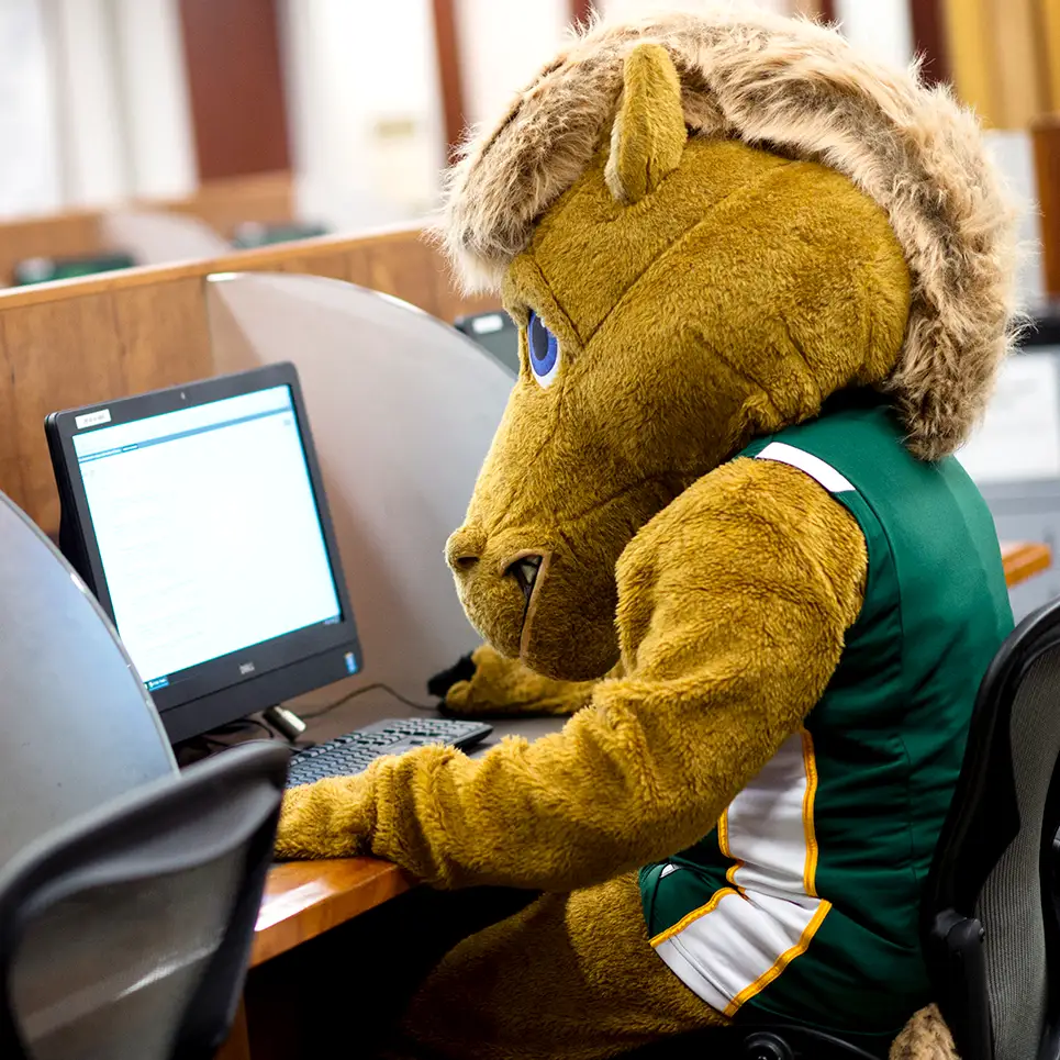 Skidmore College's horse mascot, Skids, sits at a computer station in a campus lab or library, fully suited in a green and yellow jersey, focused on the screen while typing on a keyboard.