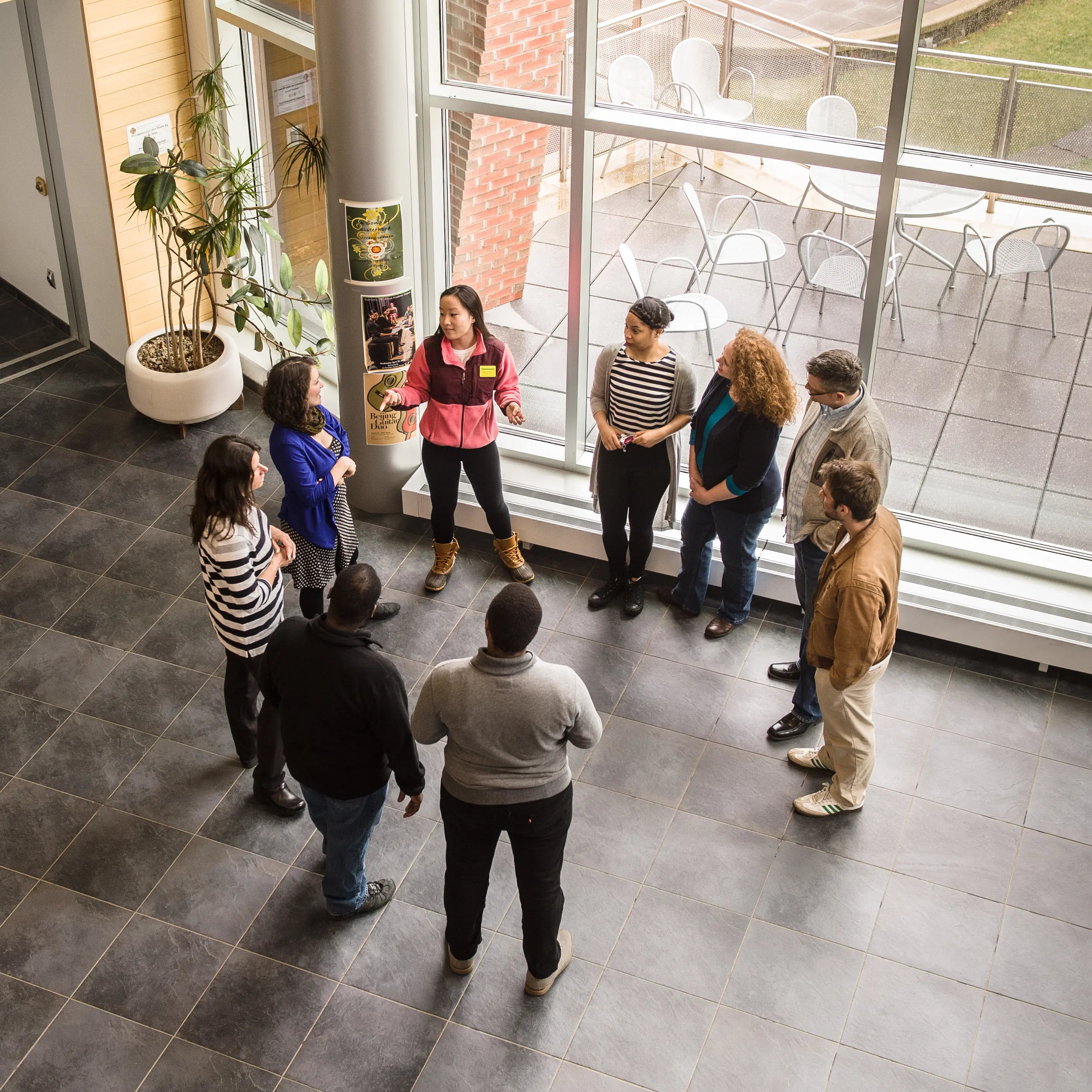 A student tour guide stands in a sunlit campus building, speaking to a small group of prospective students and families arranged in a semi-circle.