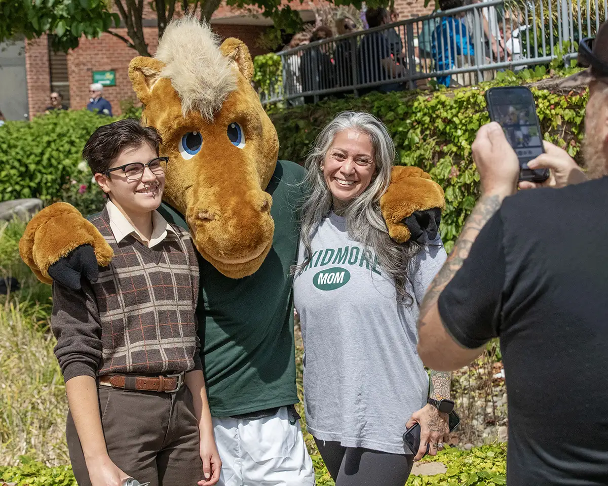 Two people posing and smiling with a Skidmore horse mascot outdoors while another person takes their photo on a smartphone, with greenery and campus buildings in the background.