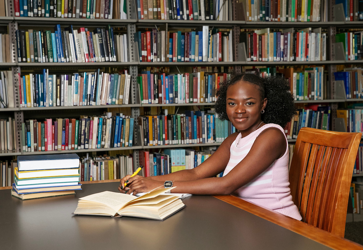 Student sits in the library at a desk with a book open and with book stacks behind her.