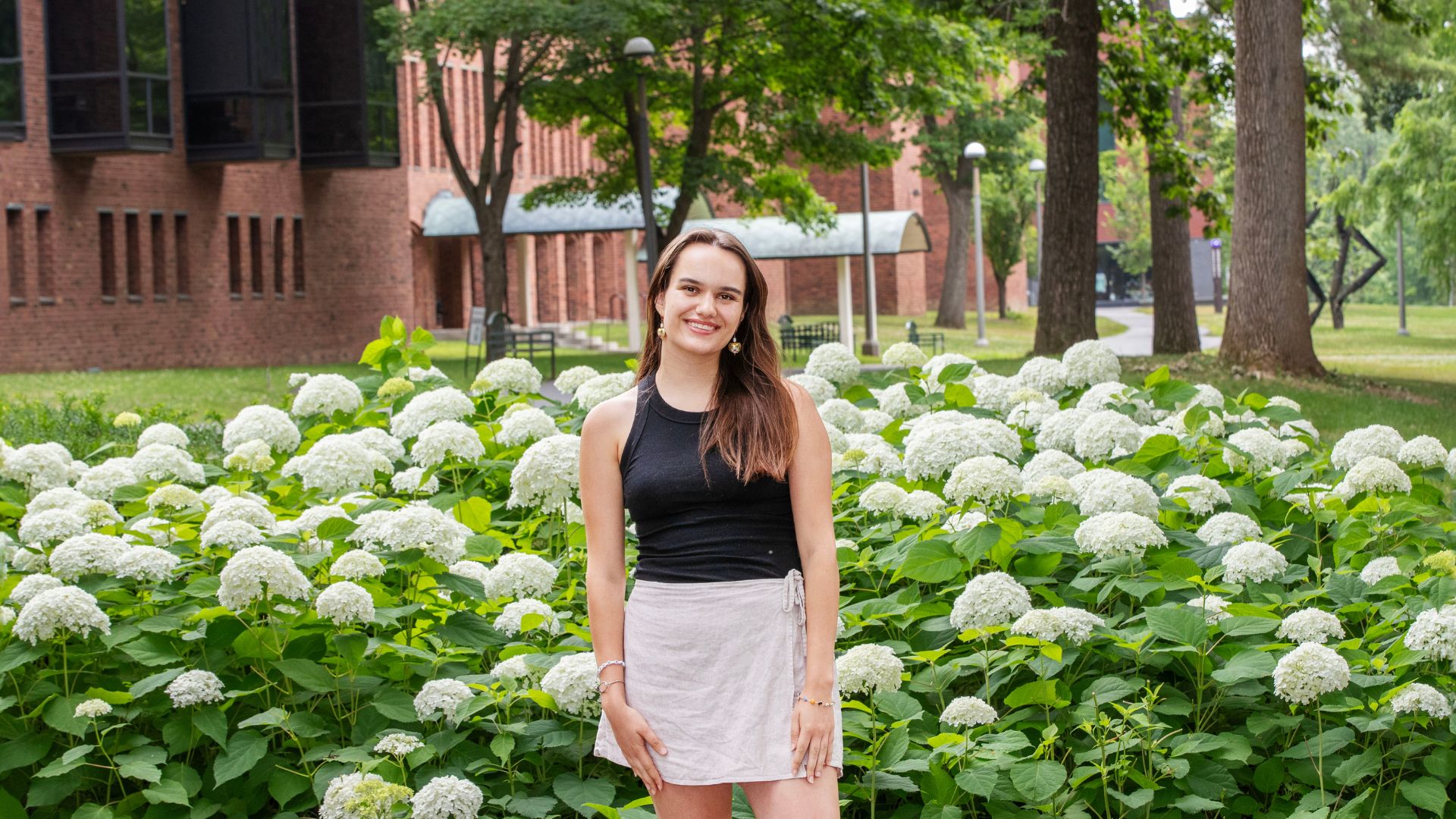 A young woman stands smiling in front of a lush garden filled with blooming white hydrangeas. She is wearing a black sleeveless top and a light beige wrap skirt. Behind her, red brick buildings and trees are visible