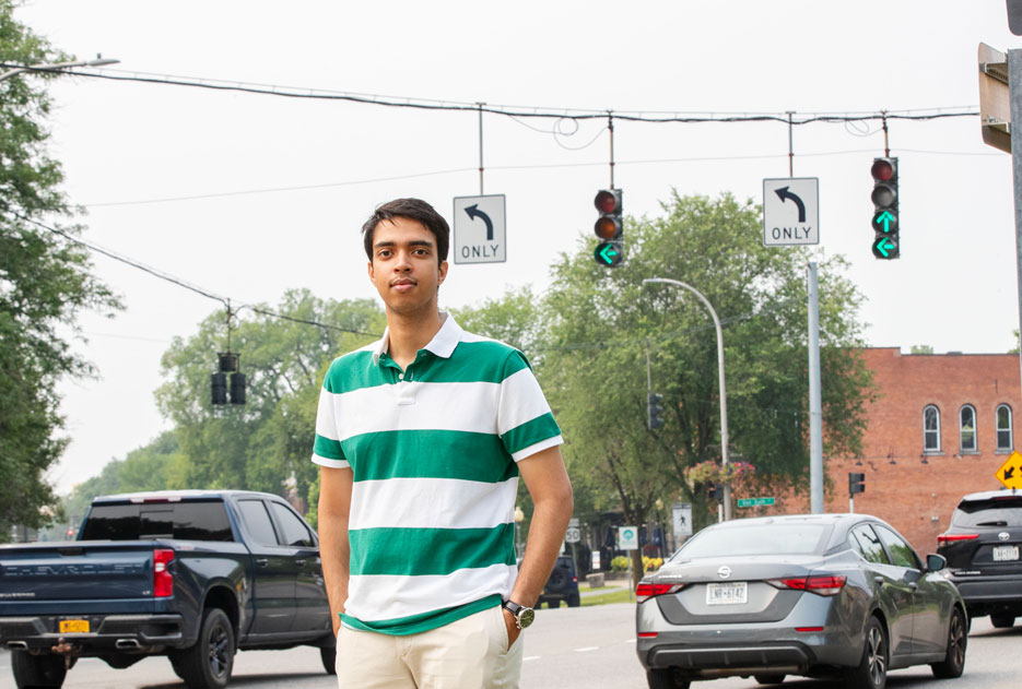Skidmore student Azizul Hakim stands in front of a traffic light as cars travel by in the distance. 