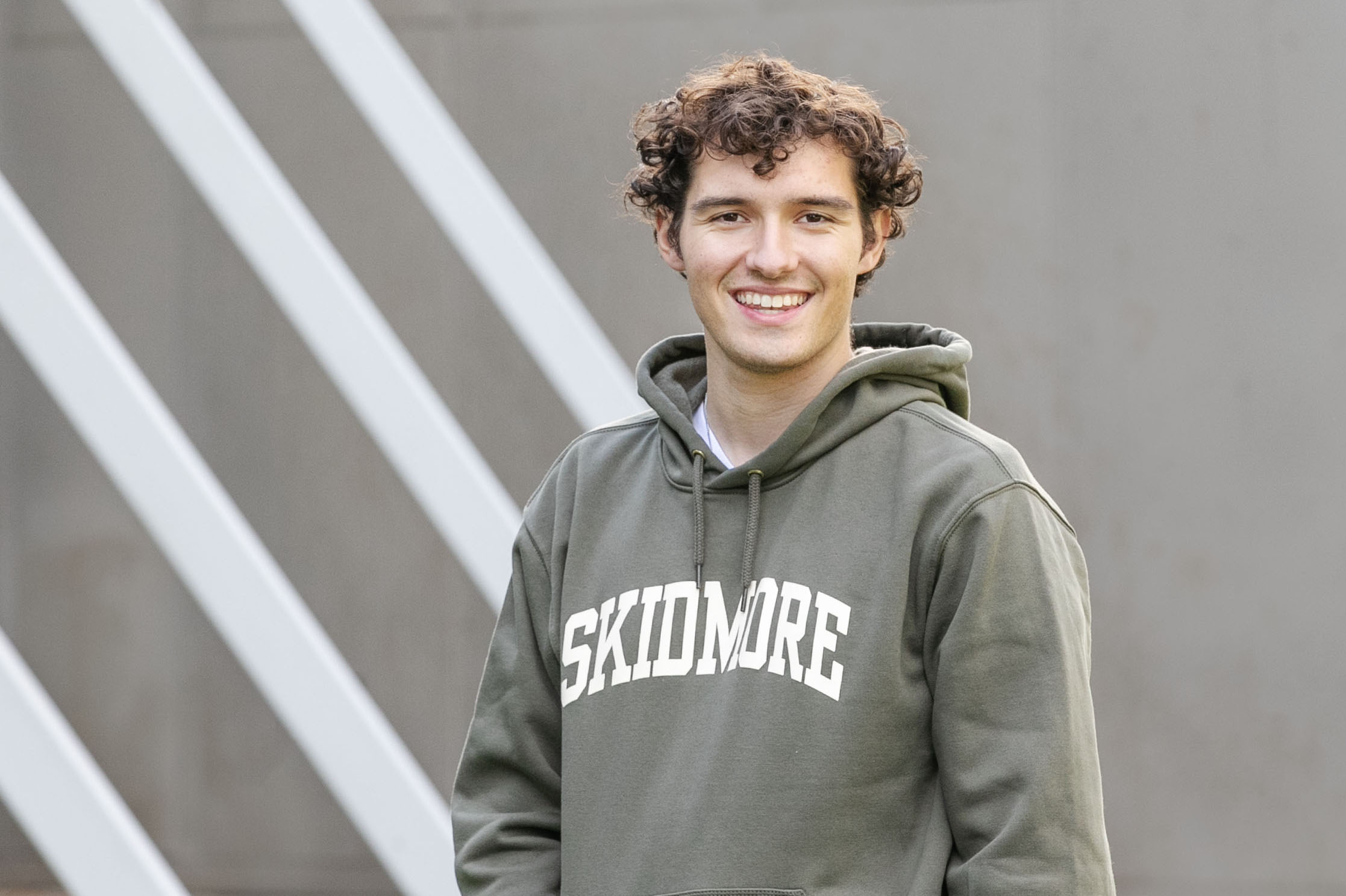 Braedon Quinlan ’24 smiles at the camera while wearing a Skidmore sweatshirt in front of the Tang Teaching Museum.