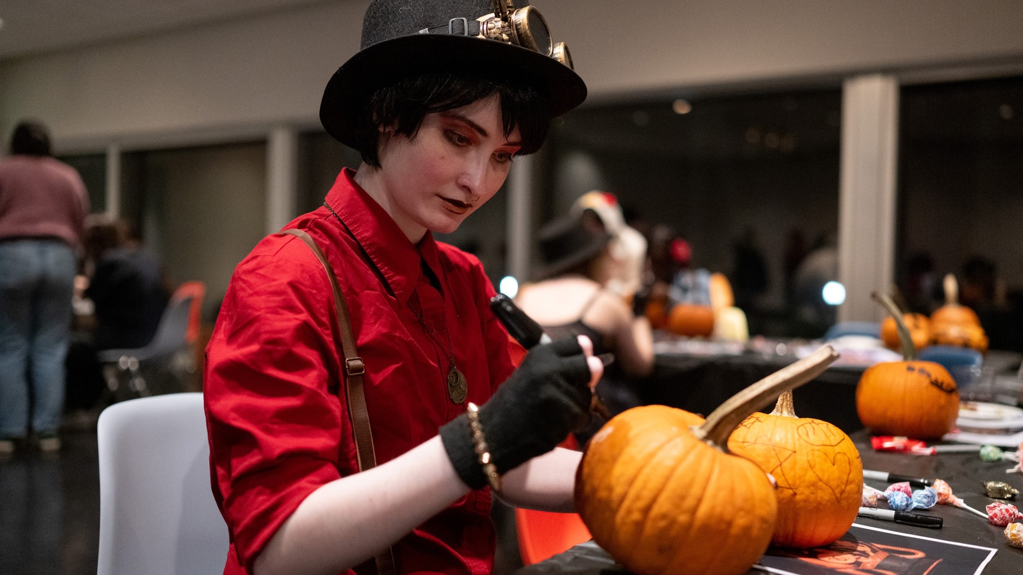 A student in an outfit that evokes the steampunk aesthetic decorates pumpkins at a table in the Tang.