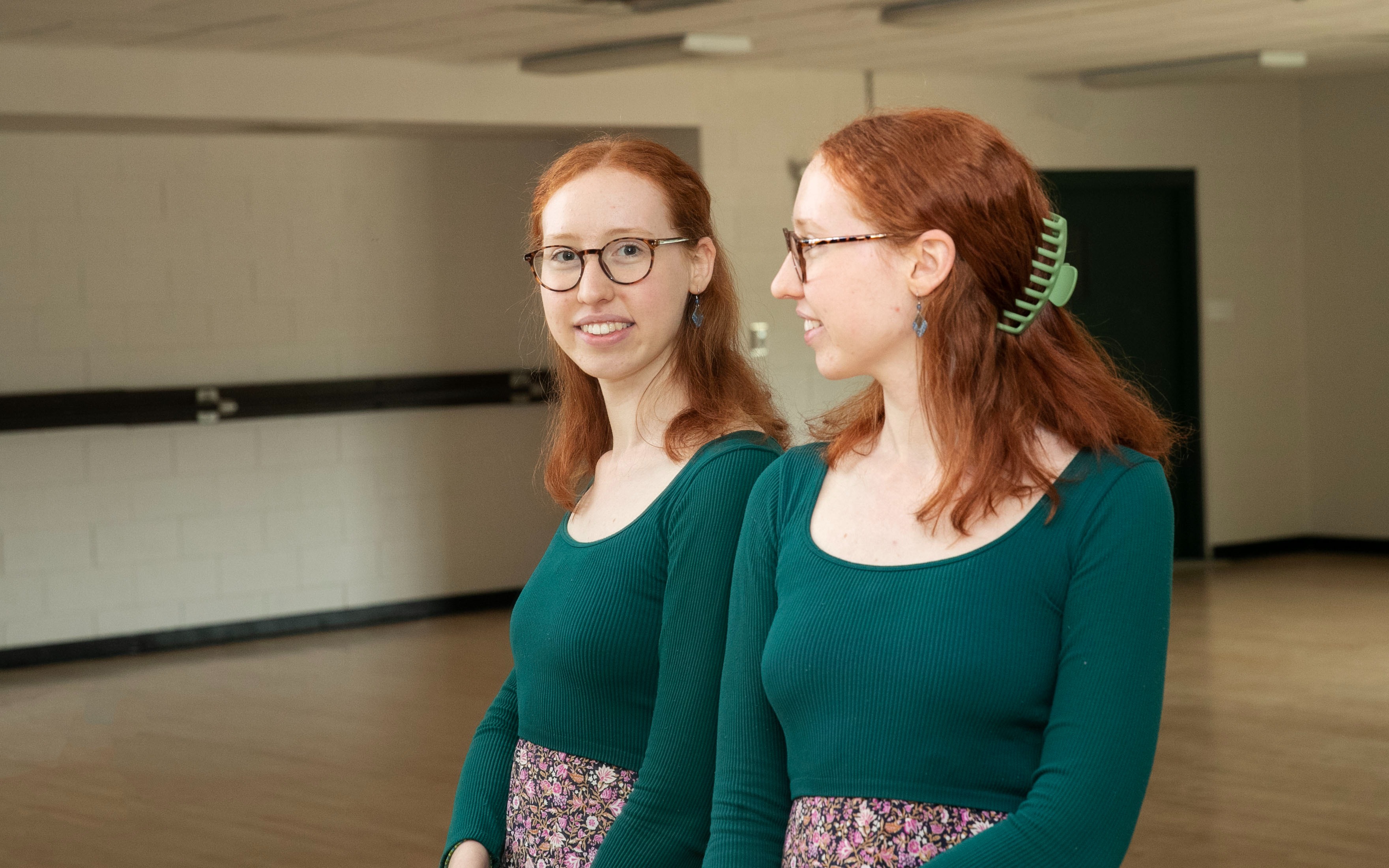 Liz Bracht '24 smiles into a mirror in a dance studio. She is wearing a blue, long sleeved top and a floral skirt.