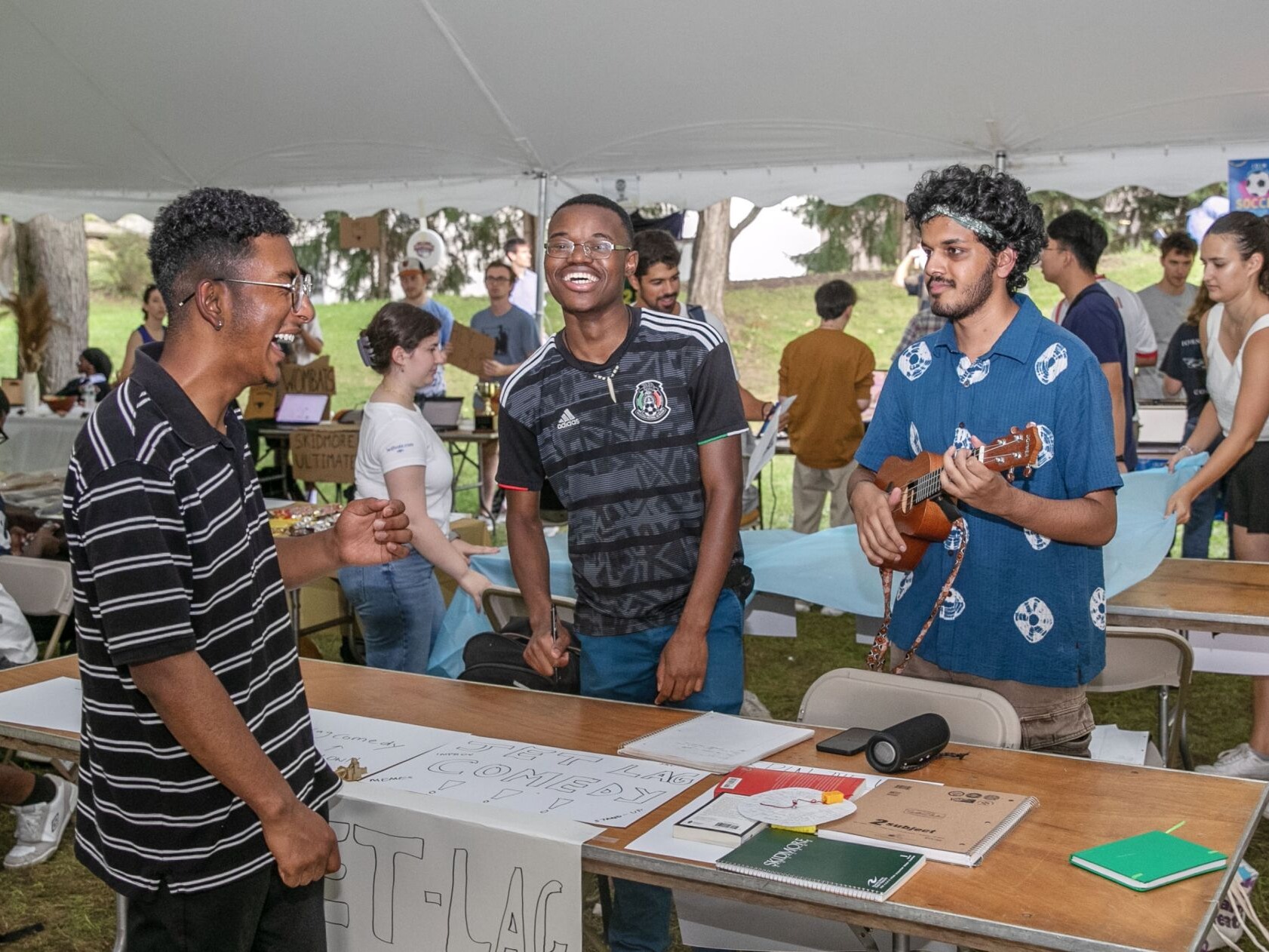 Two students laugh from a table emblazoned with "Jet Lag Comedy." Another student plays the ukulele.