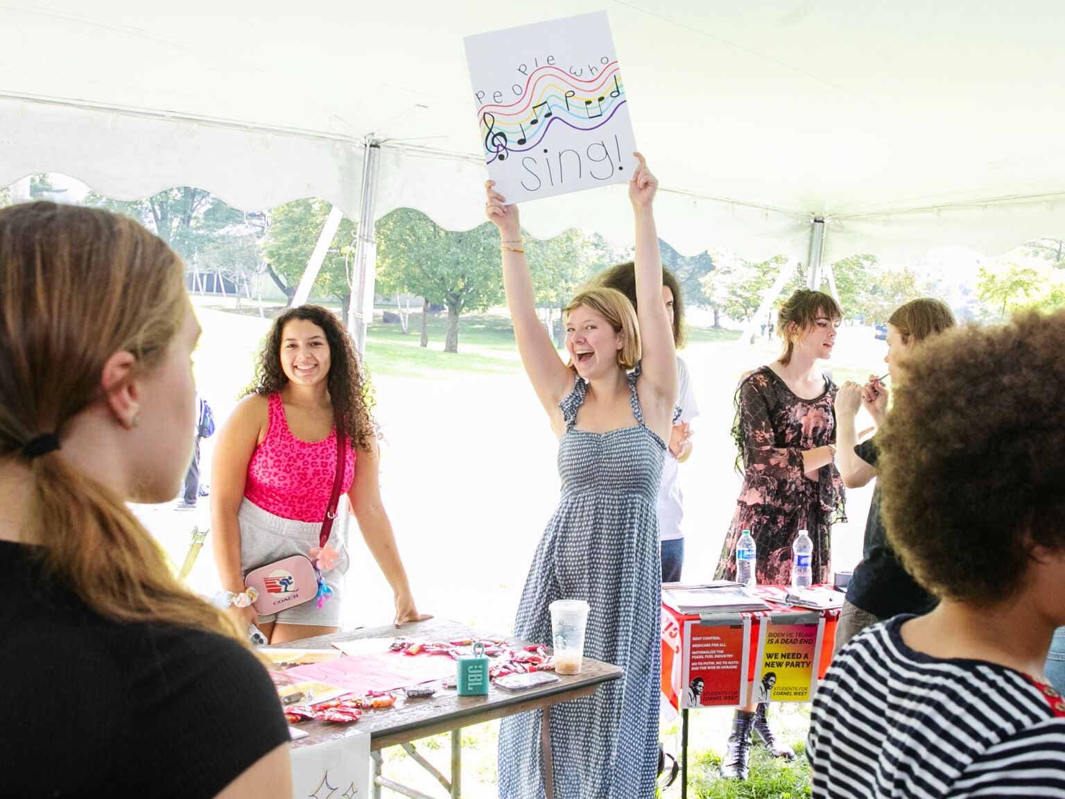 A student smiles and holds up a sign for the club. It is decorated with music notes.