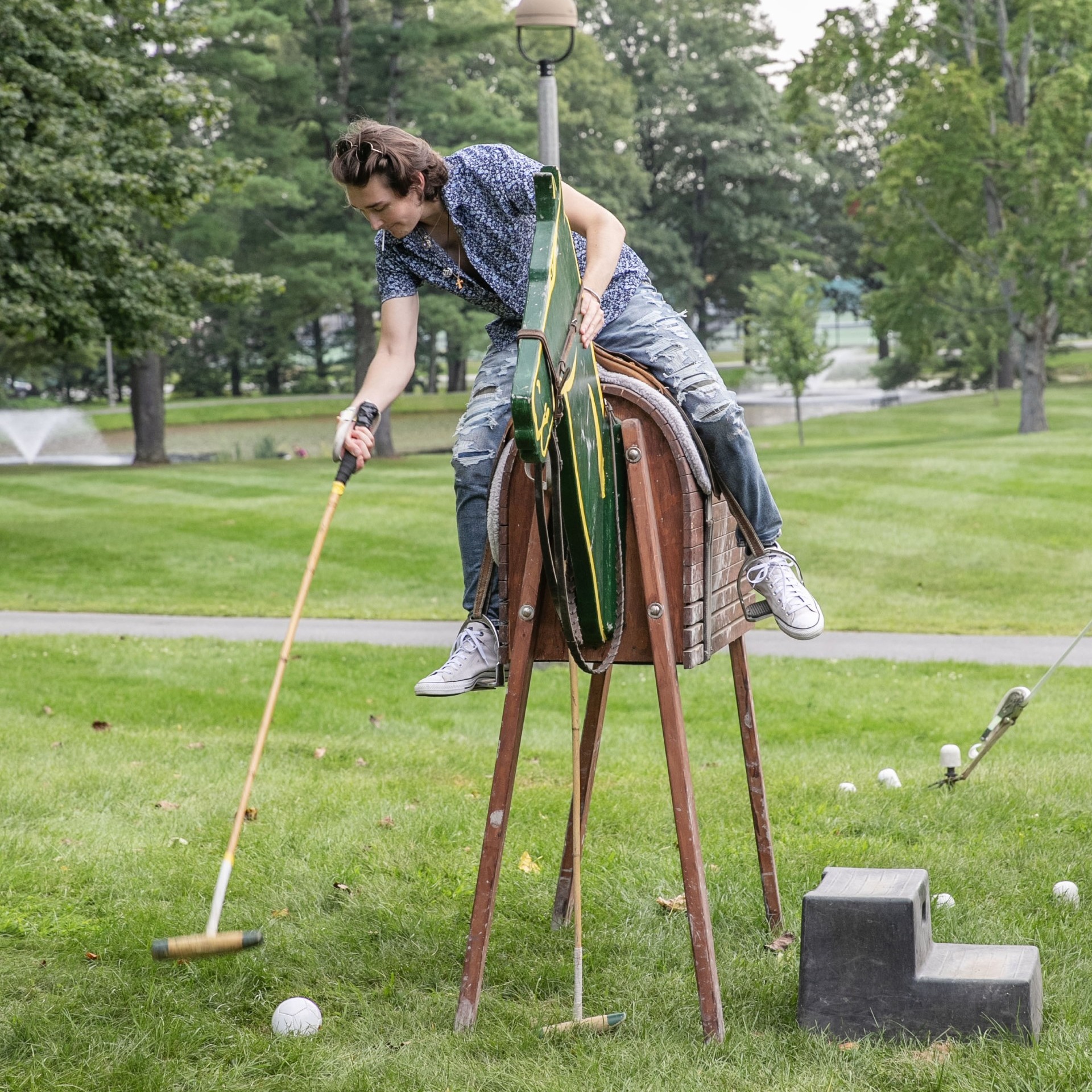 A student sits on a fake horse and reaches down to hit a polo ball with a mallet. 