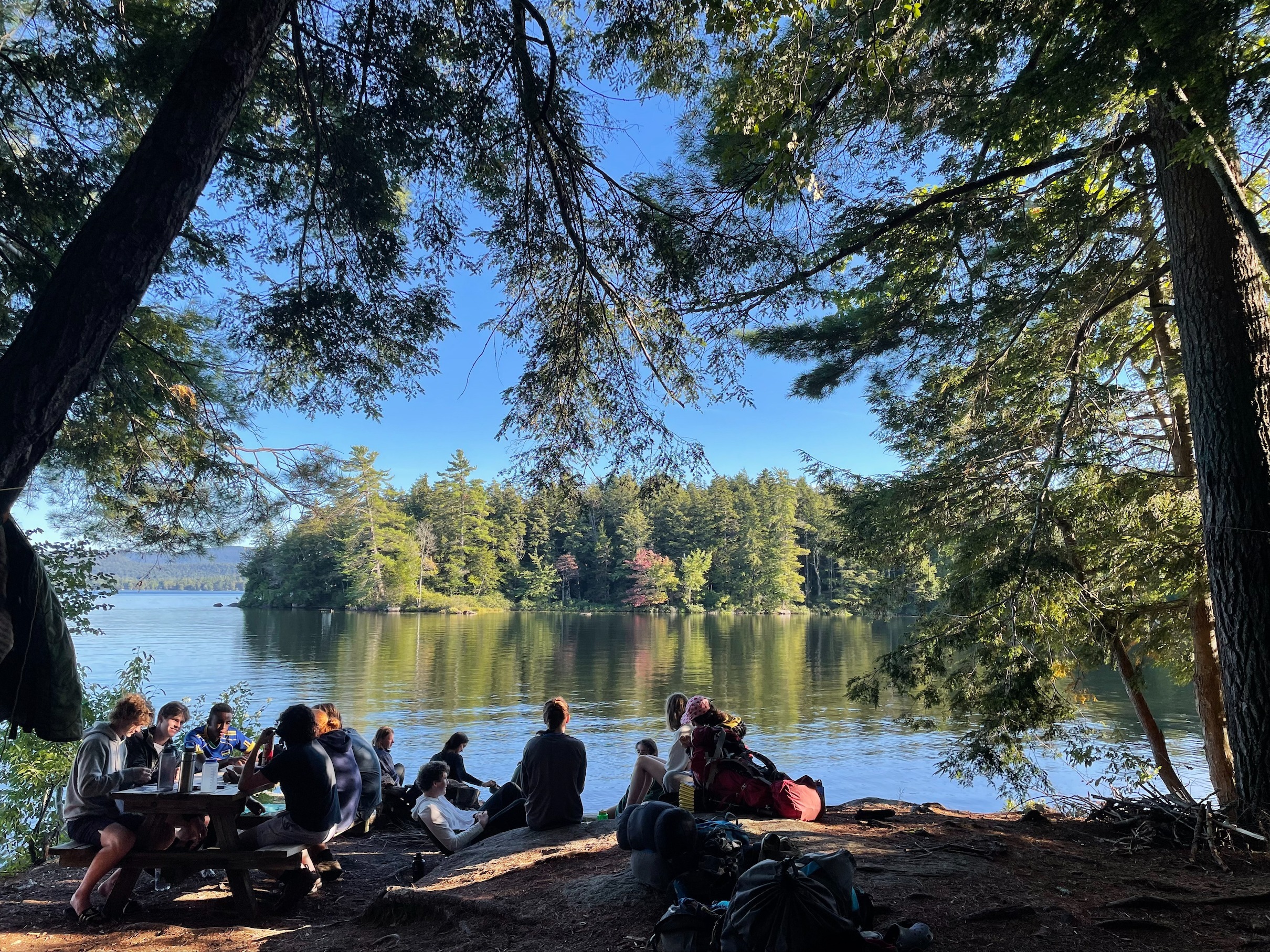 We see the backs of a group of students as they chat at a picnic table and look out across a lake.