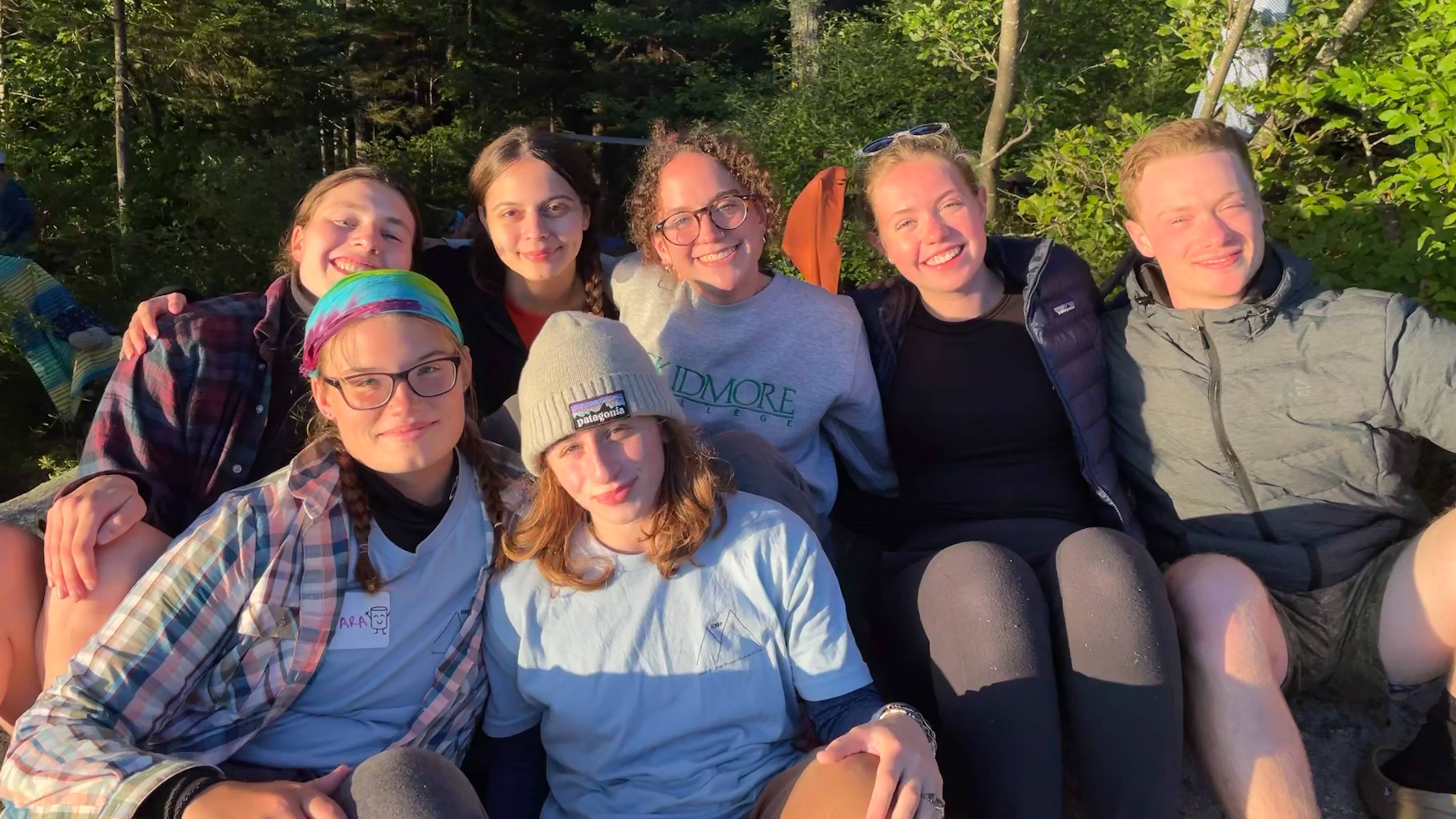 A group of seven students (including Catie Hamilton) sit and smile into the sun. Dense trees and greenery can be seen in the background.