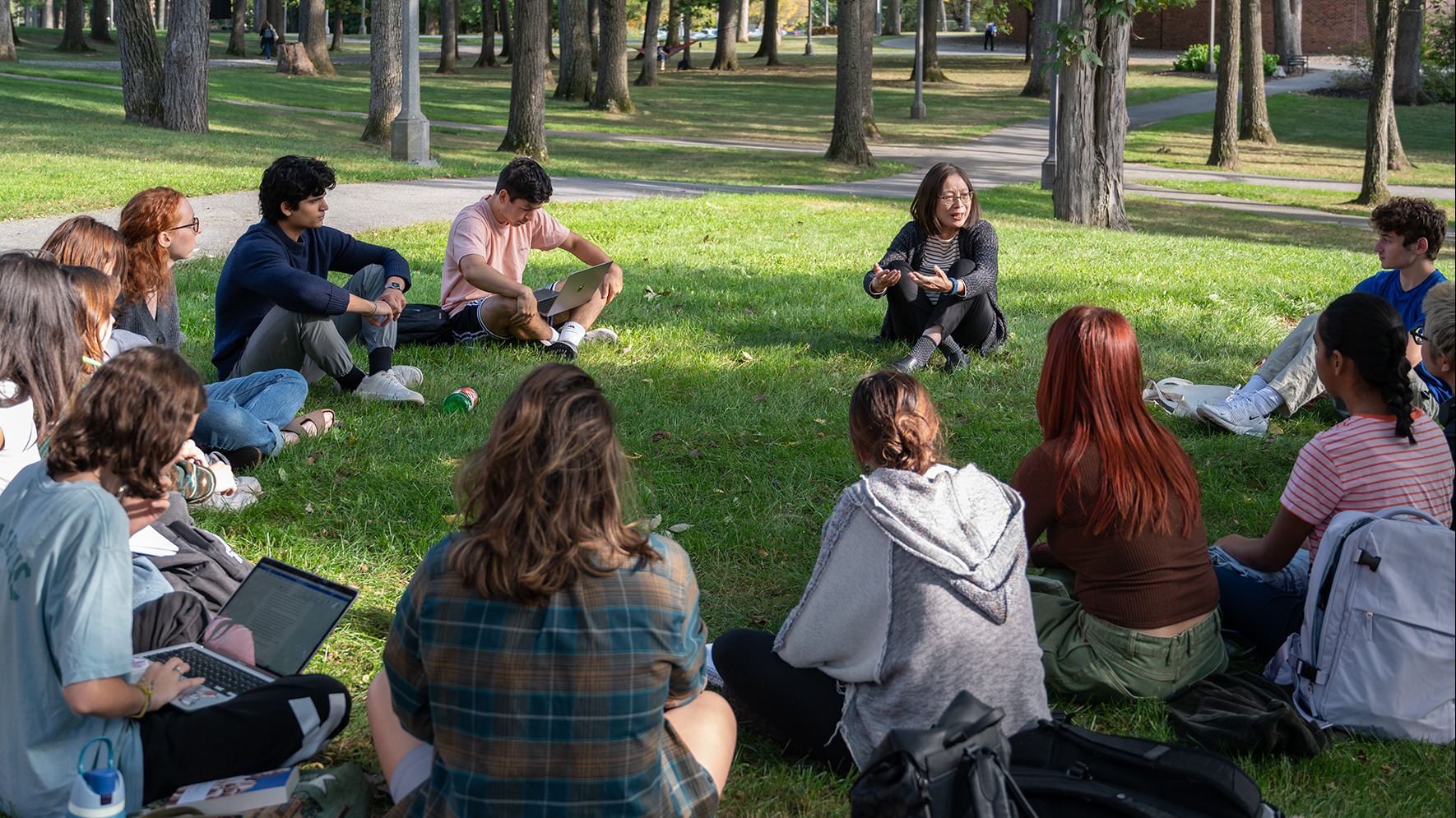 Professor Mao Chen sits in a circle on the Skidmore green with her Ideal Worlds students. They listen, engaged, as she speaks.