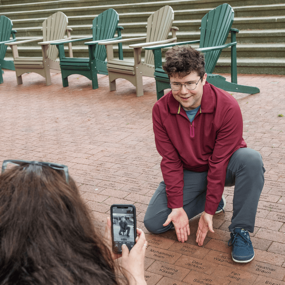 Alumni poses with his named brick, while another alum takes his photo with their phone.