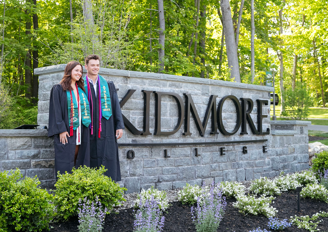 two college graduates post in front of a campus entrance sign that says Skidmore College