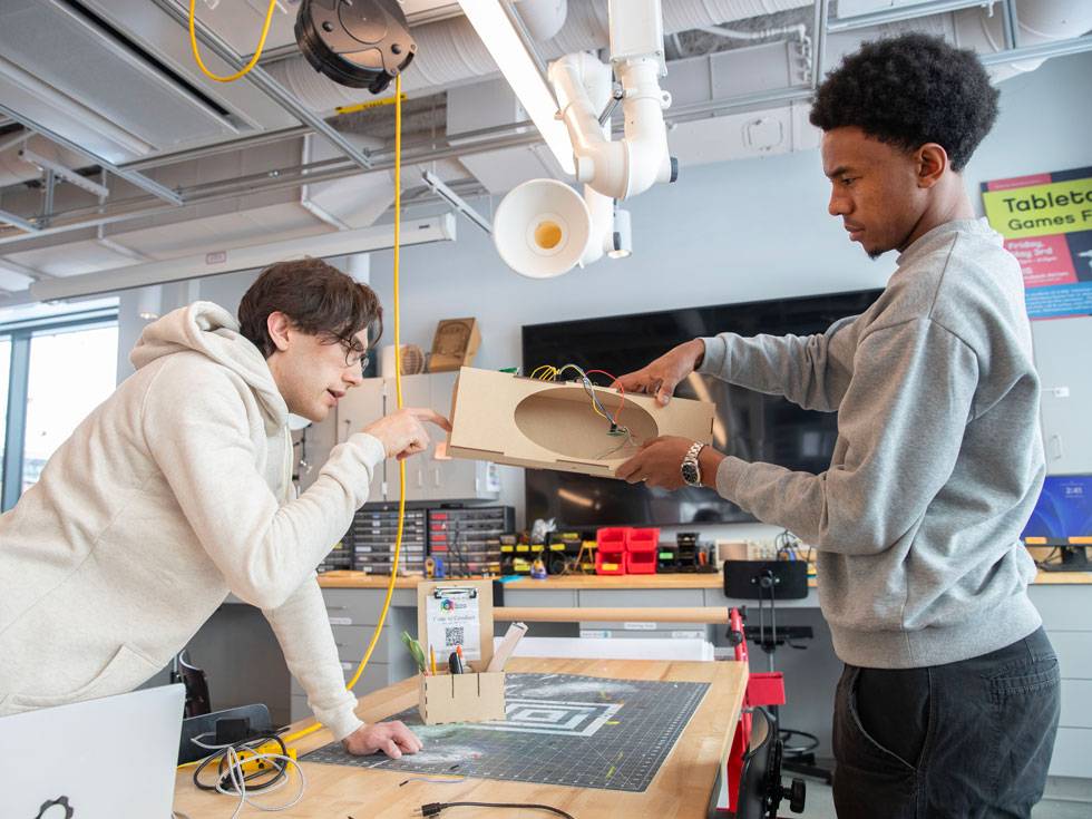 Two students work on building a project in Skidmore's ideaLab. One is holding the object over a table while they other pinpoints an area they are focused on.