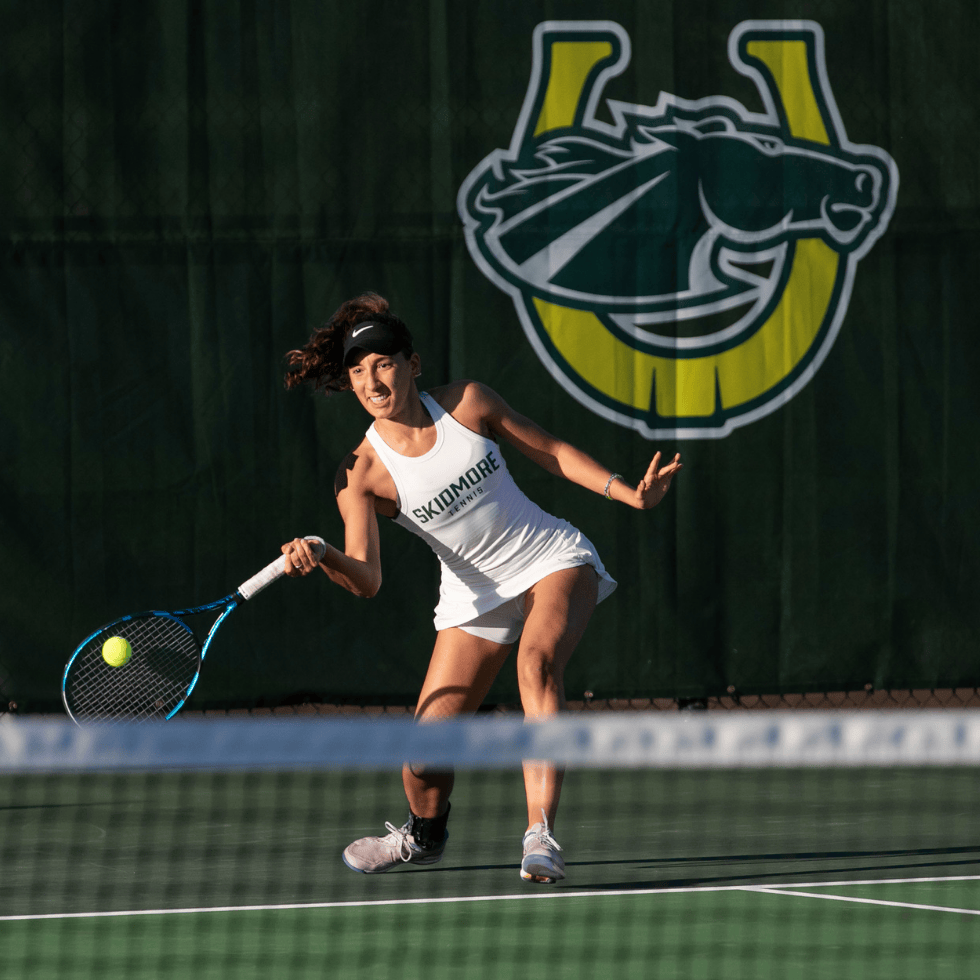 Skidmore tennis player swings at a ball on the courts with a thoroughbred logo in the background.