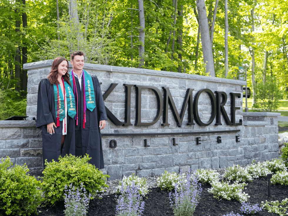 Two students standing in front of Skidmore's entrance sign wearing their graduation gowns and smiling.