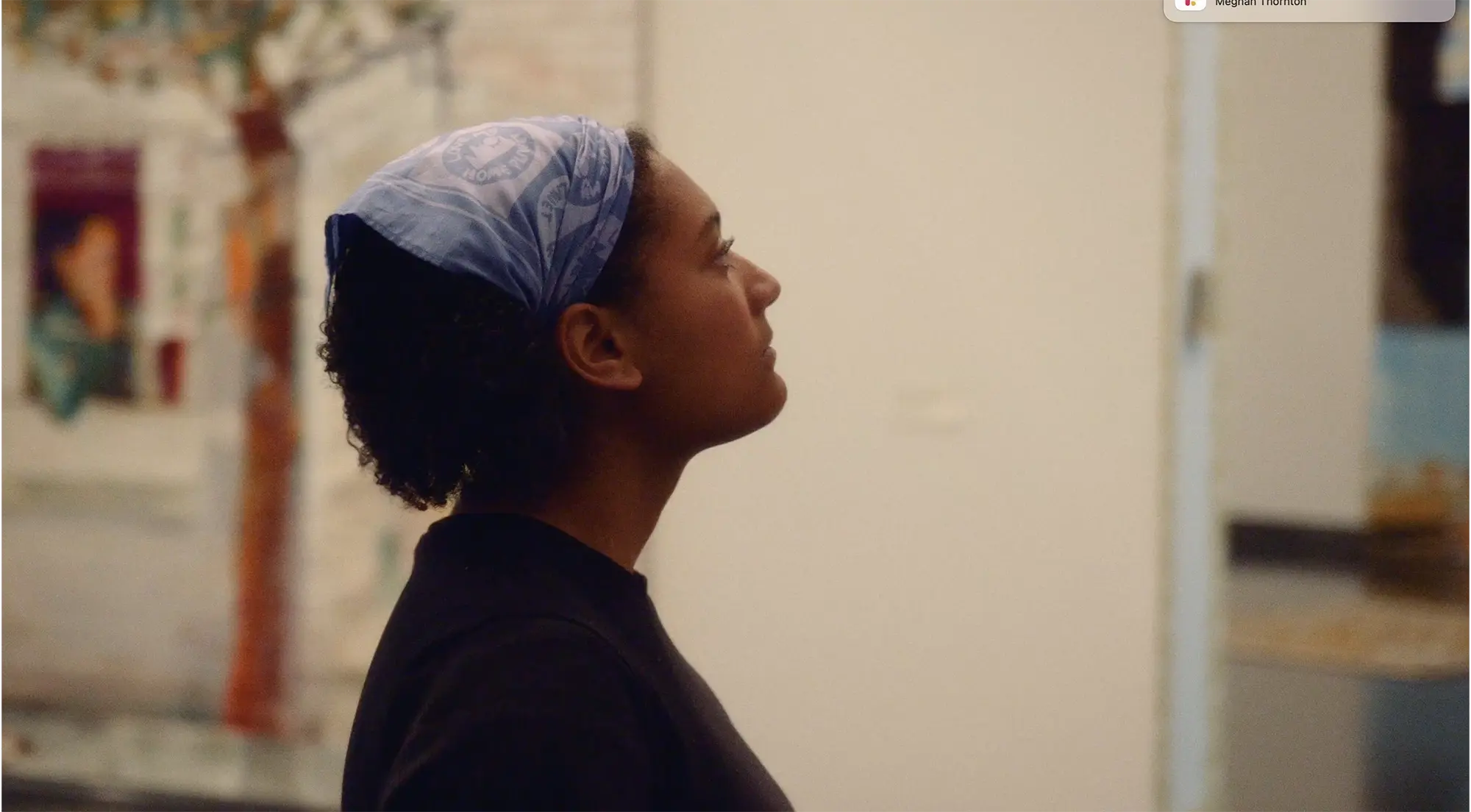 Student wearing a blue headscarf looks thoughtfully at artwork in a gallery space, with blurred paintings in the background.