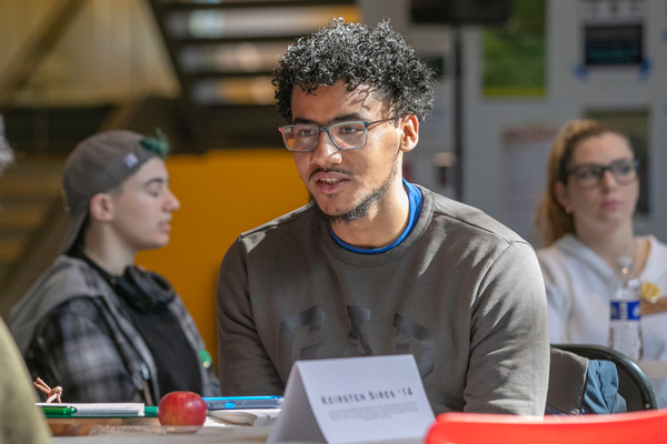 male college student sits at a table in discussion with another student