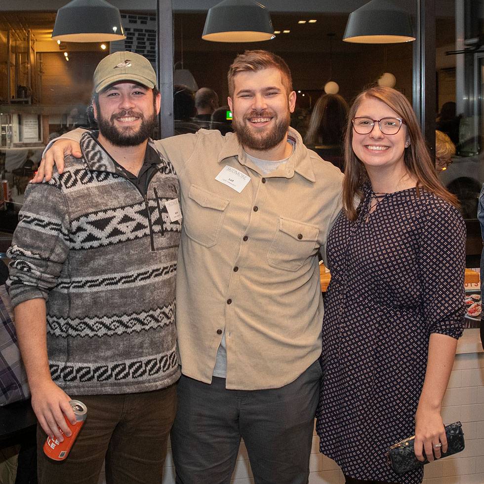 Three alumni stand smiling with their arms around one another