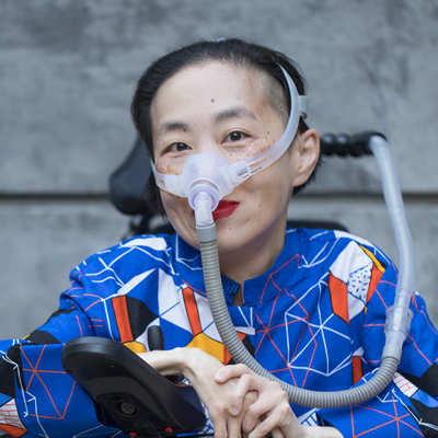 Photo of an Asian American woman in a power chair. She is wearing a blue shirt with a geometric pattern with orange, black, white, and yellow lines and cubes. She is wearing a mask over her nose attached to a gray tube and bright red lip color. She is smiling at the camera. Photo credit: Eddie Hernandez Photography
