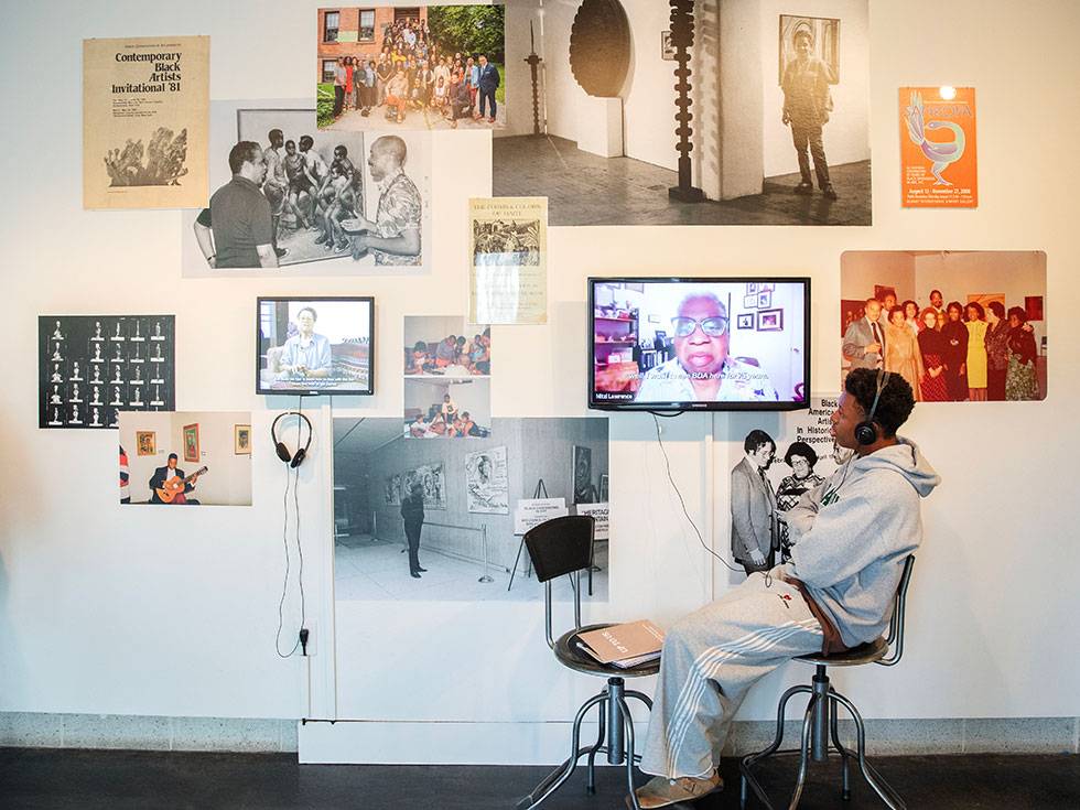 A student sits in a chair wearing headphones, listening to a video that is part of an art exhibit at the Tang Teacher Museum and Art Gallery. Not only is there a tv with a video paying, but also a gallery wall of different images.