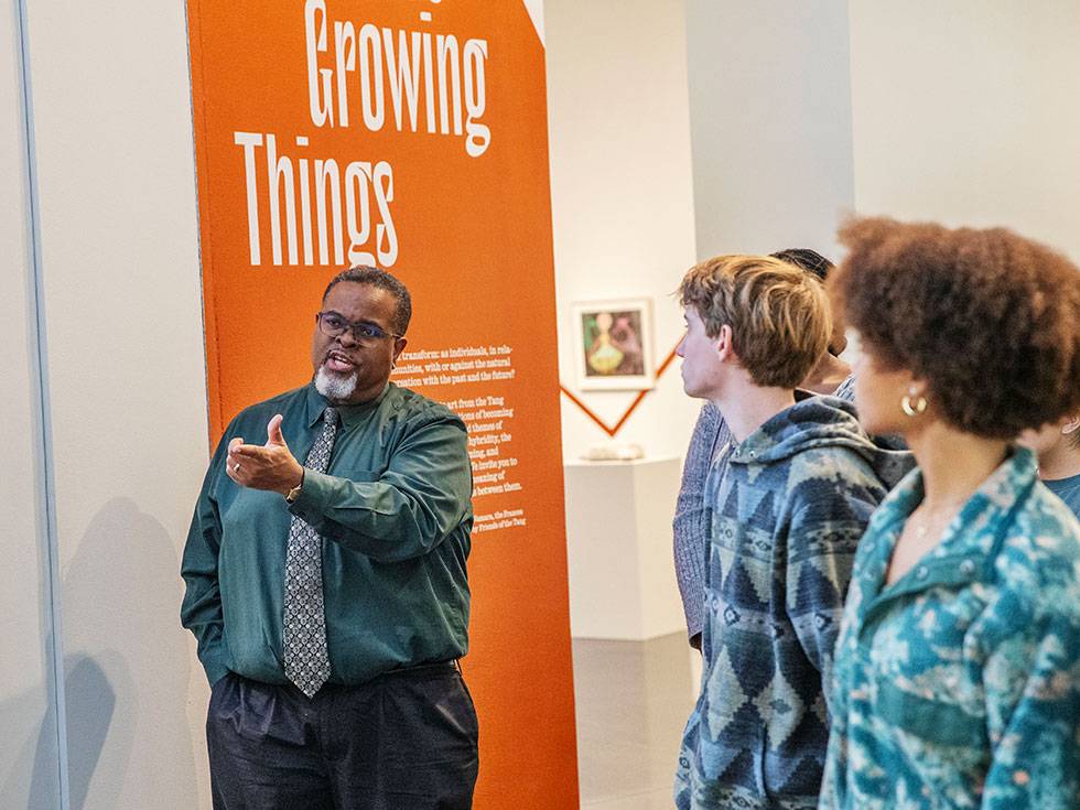 Professor Winston Grady-Willis stands while holding out a hand to gesture towards a subject he and students are examining in an exhibit in the Tang Teaching Museum and Art Gallery. He is wearng a green button down shirt with a tie, and there is an orange poster behind him with white text that reads "Growing Things"