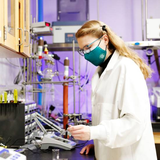 A scientist wearing a white lab coat and a dark green face mask works in a chemistry laboratory, using lab equipment and instruments, surrounded by glassware and tubes.