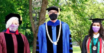 President Glotzbach, Jennifer Mueller, and Jinan Al-Busaidi stand for a photo with graduation outfits and masks