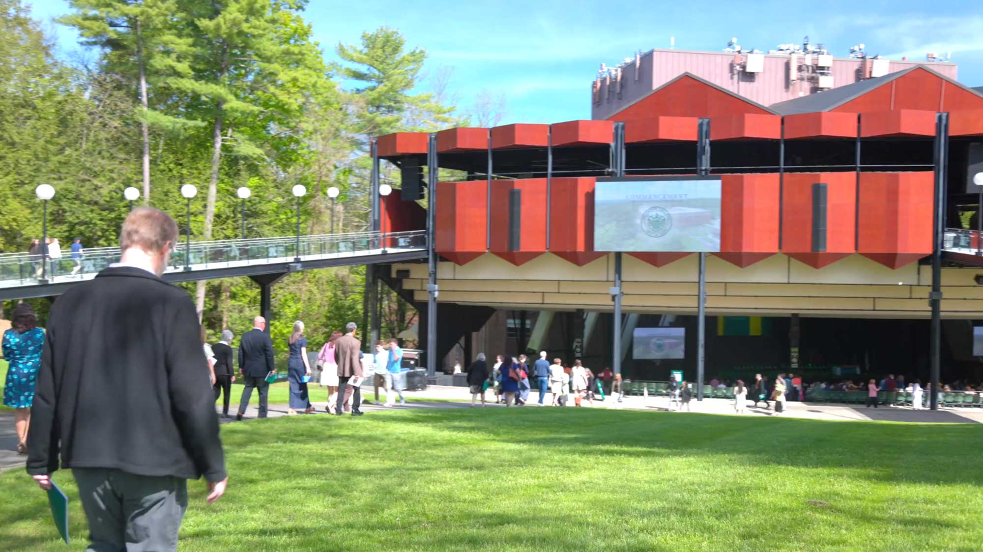 Commencement guests walk across a sunny lawn toward the ceremony venue on a bright spring day.