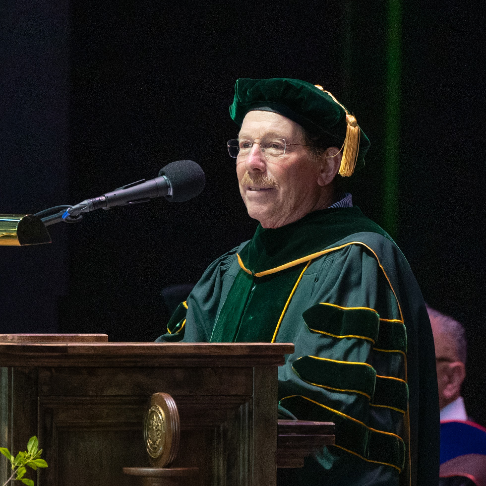 Jon P. Achenbaum ’77 in green academic regalia speaks at a podium with faculty seated behind him.